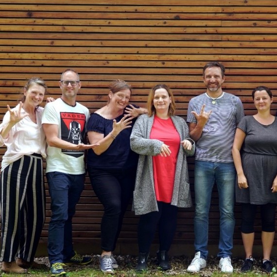 Sechs Personen stehen vor einer Wand und zeigen Geb&auml;rden.