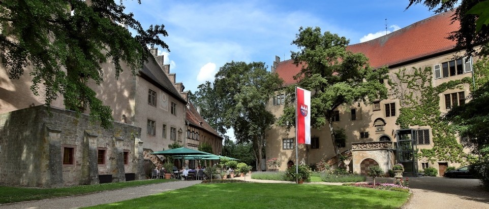Um einen begr&uuml;nten Hof gruppieren sich zwei historische Schlossgeb&auml;ude. Das Hauptschloss ist dreist&ouml;ckig. Vor dem kleineren Schloss stehen sind Sonnenschirme, St&uuml;hle und Tische.