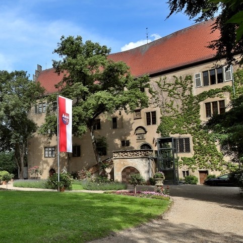 Ein dreist&ouml;ckiges Schlossgeb&auml;ude. Davor steht ein gro&szlig;er Baum und eine rot-wei&szlig;e Fahne.