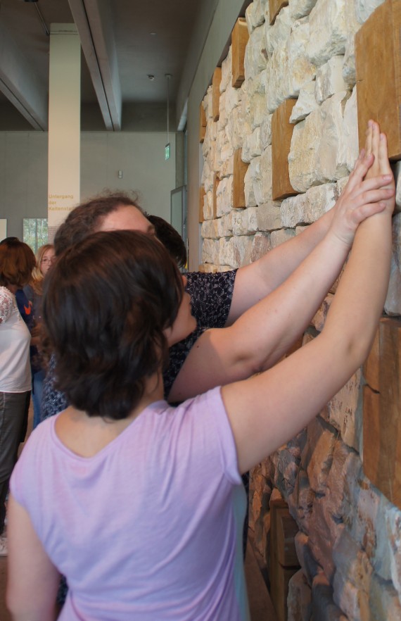 Zwei Personen stehen vor einer Steinwand. Sie fassen die Wand an. Einer Person f&uuml;hrt dabei die andere Person.