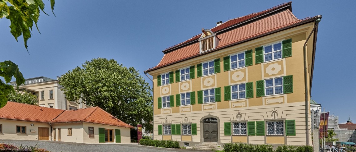 Dreist&ouml;ckiges Geb&auml;ude mit Walmdach. Die Fenster sind gleichm&auml;&szlig;ig verteilt und von gr&uuml;nen Fensterl&auml;den umrahmt.