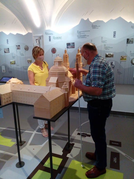 Eine Frau und ein Mann stehen neben einem Tastmodell einer Basilika. Der Mann h&auml;lt einen Blindenstock in der einen Hand. Mit der anderen Hand ber&uuml;hrt er das Modell.