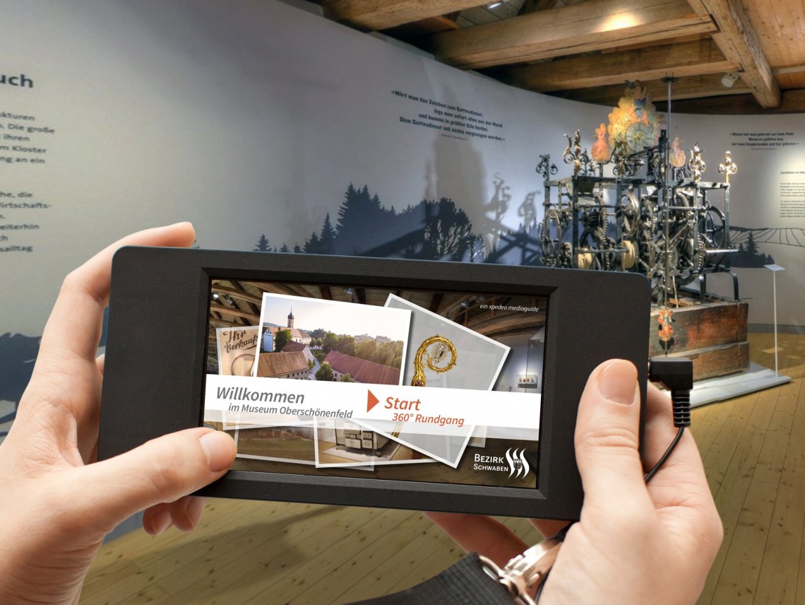 zwei H&auml;nde halten einen Medienguide. Im Hintergrund ist ein Ausstellungsraum zu sehen.