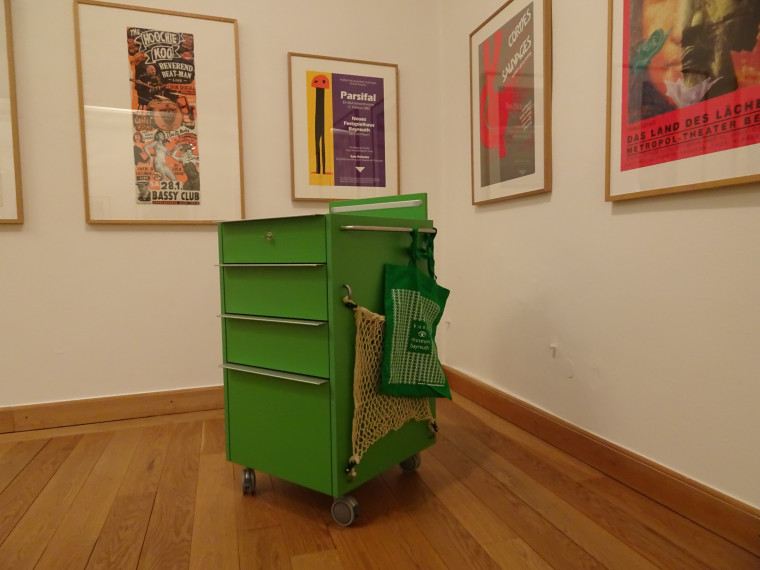 Ein gr&uuml;ner Rollcontainer vor gerahmten Postern an einer Wand.