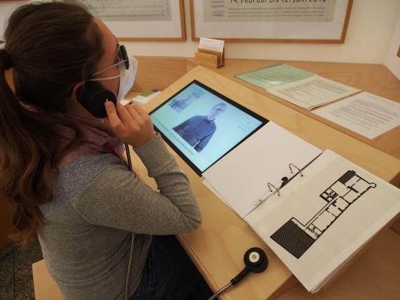 Eine Frau h&auml;lt sich an einer Medienstation einen H&ouml;rer an das Ohr. Auf dem Bildschirm vor ihr l&auml;uft ein Film ab.