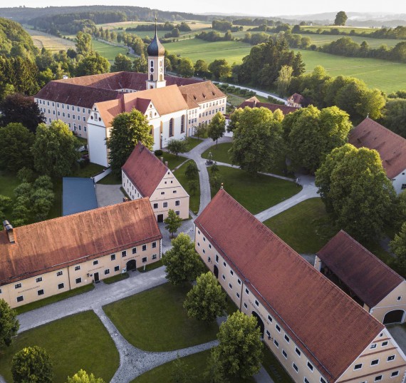 Ein Klostergeb&auml;ude mit Kirche und Zwiebelturm steht in einer Landschaft mit Wiesen und Hecken.