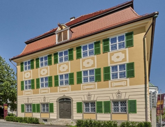 Dreist&ouml;ckiges Geb&auml;ude mit Walmdach. Die Fenster sind gleichm&auml;&szlig;ig verteilt und von gr&uuml;nen Fensterl&auml;den umrahmt.