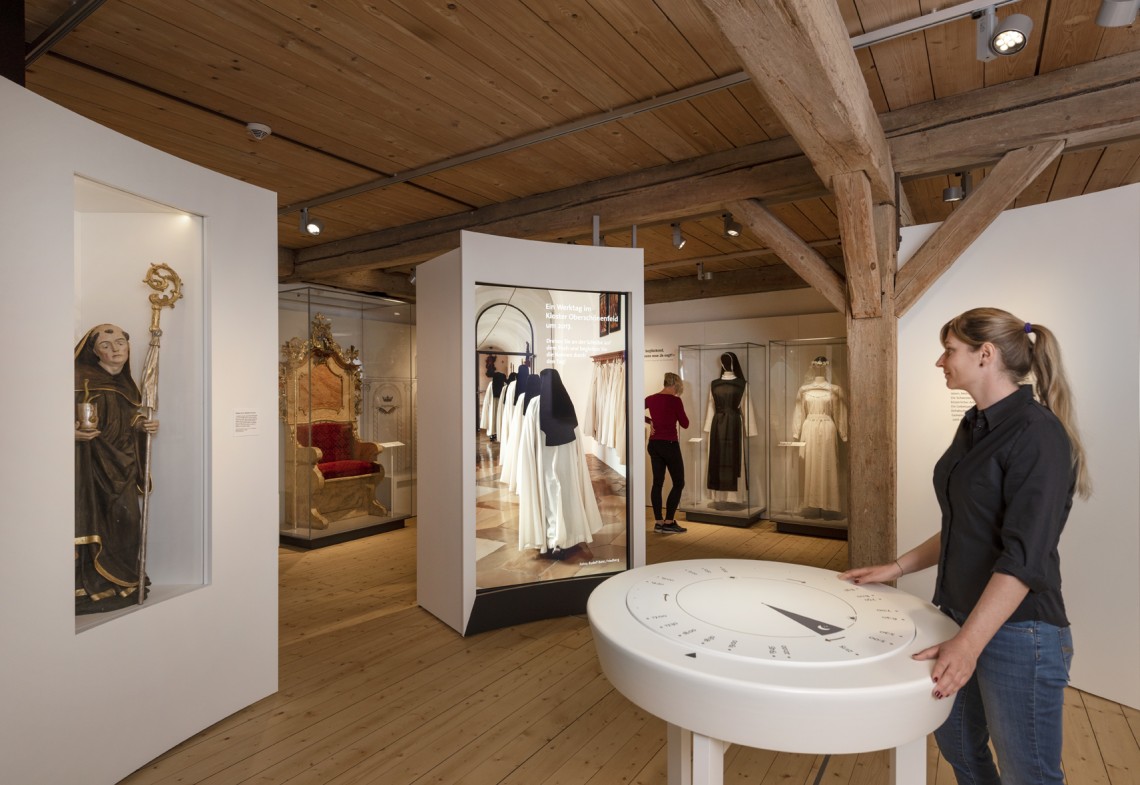 Eine Frau steht an einem runden Tisch in einem Ausstellungsraum mit vielen Vitrinen.