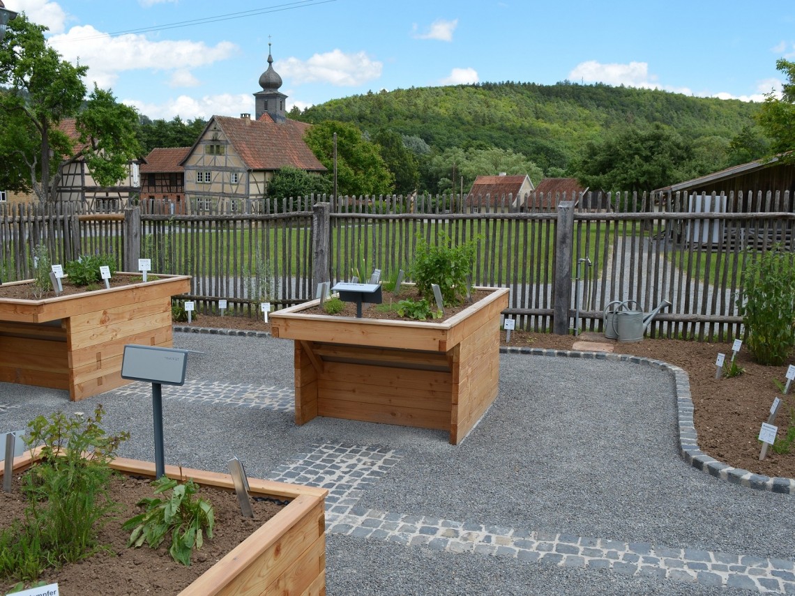 Hochbeete mit Kr&auml;utern sind von einem Zaun eingerahmt. Im Hintergrund ist ein Fachwerkhaus und ein Kirchturm mit Zwiebelhaube zu sehen.