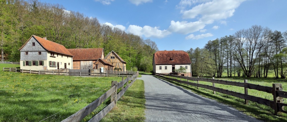 Ein geschotterter Weg f&uuml;hrt an zwei Viehweiden mit Holzzaun auf zwei historischer Geb&auml;ude hin. Eines der Geb&auml;ude ist ein Dreiseithof.