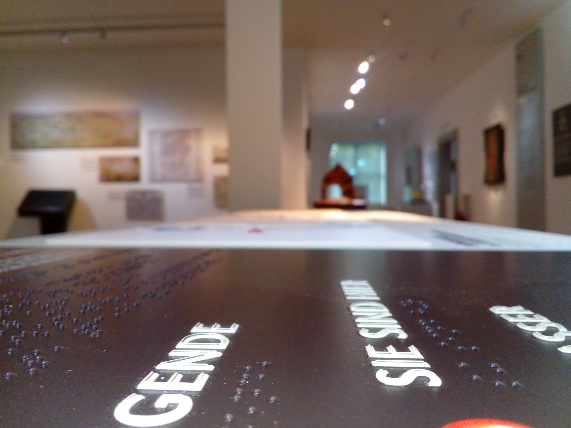 Blick in einen Ausstellungsraum, im Vordergrund ist ein Text in Brailleschrift zu erkennen.