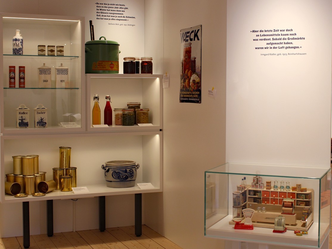 in einer Vitrinen steht ein Kaufmannsladen zum Spielen. Im Hintergrund stehen in einem Regal an der Wand Objekte.