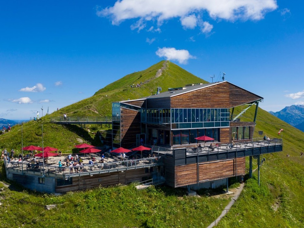 Eine Bergstation an einem begr&uuml;nten Gipfel. Auf der Terrasse stehen rote Sonnenschirme. Der Himmel ist blau.
