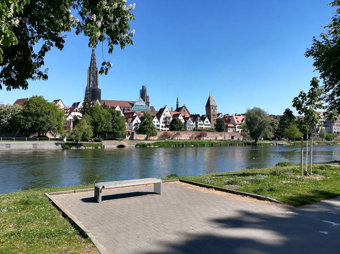 Eine Bank steht an einem Fluss. Im Hintergrund ist eine Stadt mit Kircht&uuml;rmen.
