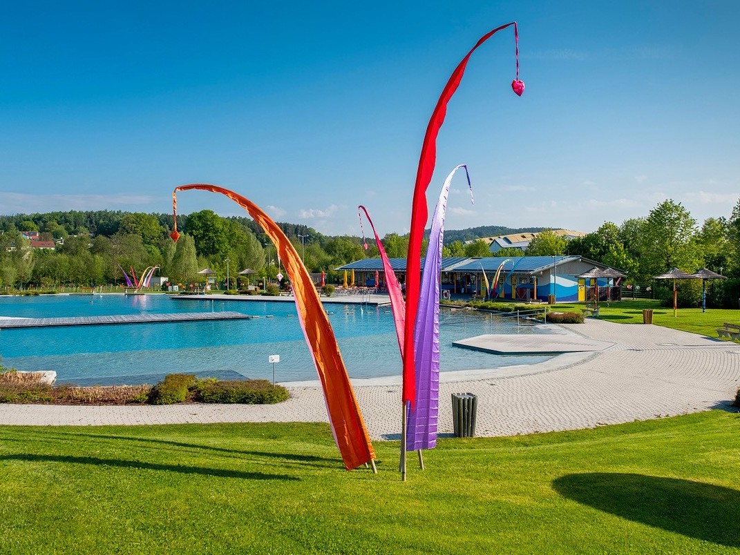 drei bunte Fahnen vor einem organisch geformten Schwimmbecken. 