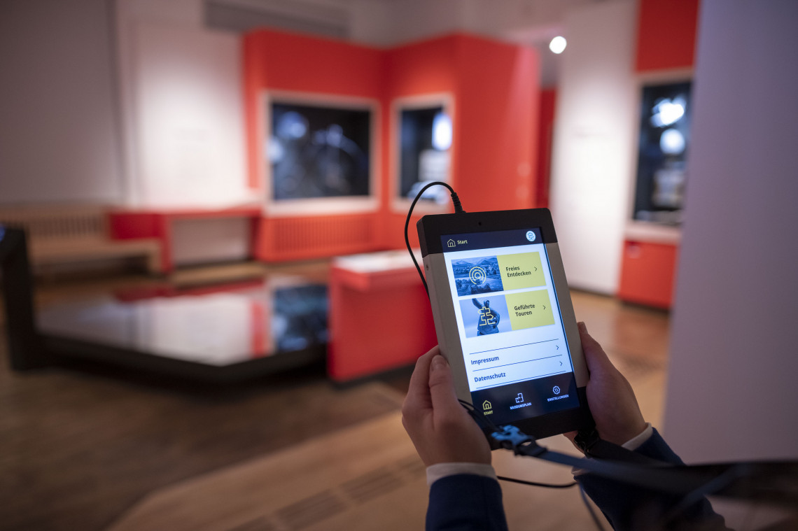 Ein Tablet mit Informationen zur Ausstellung wird von zwei H&auml;nden gehalten. Im Hintergrund ist die Ausstellung unscharf zu sehen.