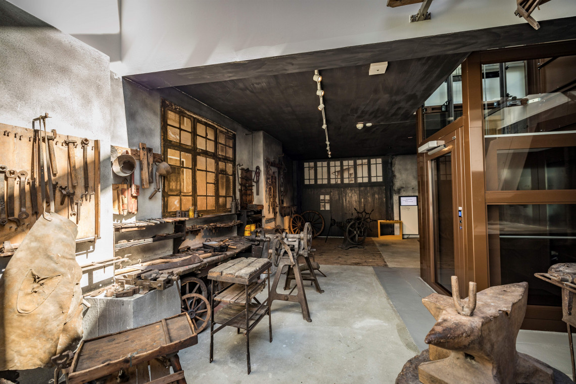 Ein Aufzug in der Raummitte f&uuml;hrt direkt in einen Ausstellungsraum, der eine historische Schmiede mit Werkzeugen an der Wand, Amboss und weitere Ger&auml;tschaften beherbergt.