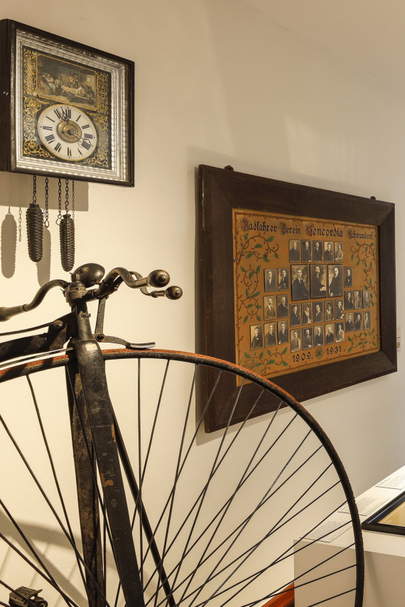 Eine historische Pendeluhr und eine historische Tafel mit Mitgliedern des Radfahrvereins h&auml;ngen an einer Wand. Davor steht ein Hochrad.