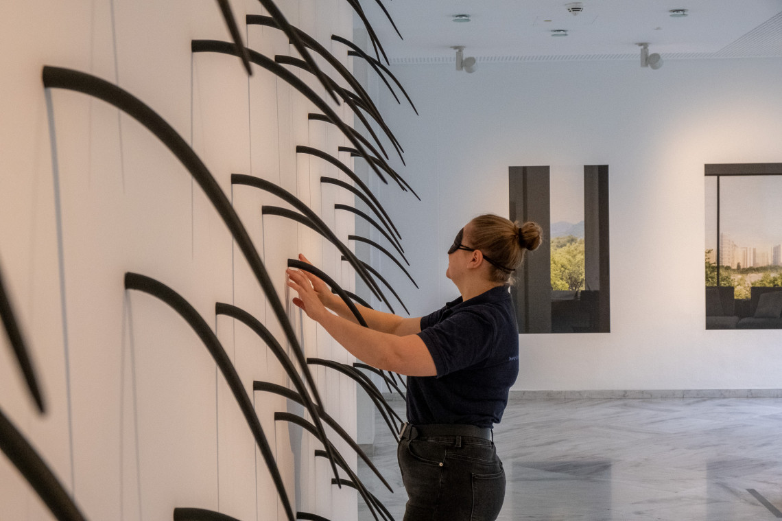 Aus einer Ausstellungswand wuchern viele schmale spitz zulaufende, nach unten h&auml;ngende Kunstobjekte der K&uuml;nstlerin Nicole Frenzel mit dem Titel "hanging around". Eine Frau mit Dunkelbrille steht davor und ertastet diese Kunstwerke.