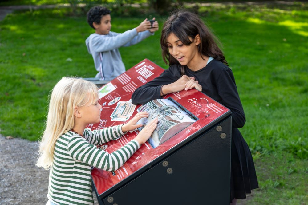 Zwei Kinder probieren eine Mitmachaktion im Park aus, ein Kind schaut sich auf dem Smartphone die begleitende Cambodunum App an
