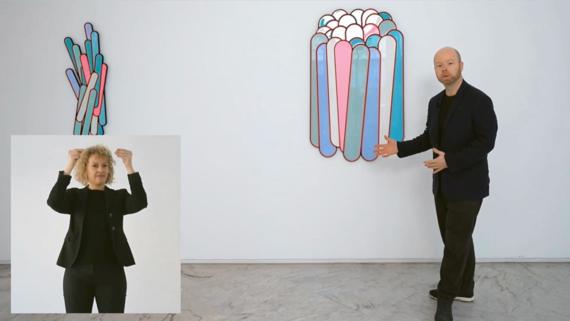 Ein Mann steht an einer Wand mit einem Kunstwerk, das aus Glasfl&auml;chen zu bestehen scheint. Im Vordergrund ist auf einem Screen eine Geb&auml;rdendolmetscherin eingeblendet.